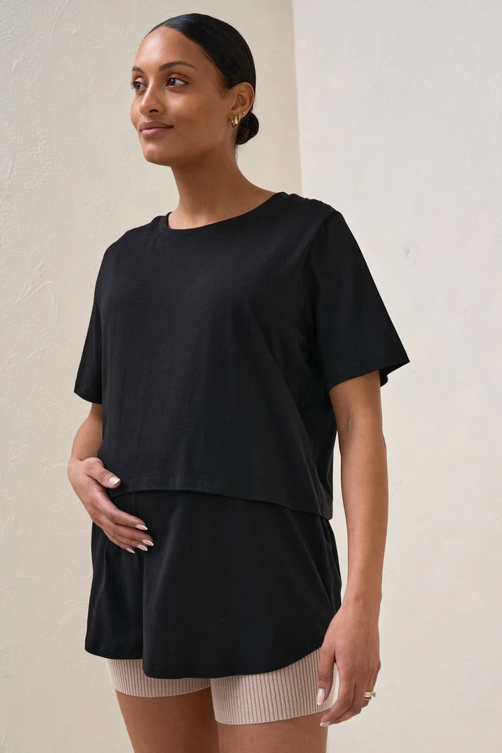 A pregnant woman wearing a black nursing tee with short sleeves and a round neck, paired with beige shorts.