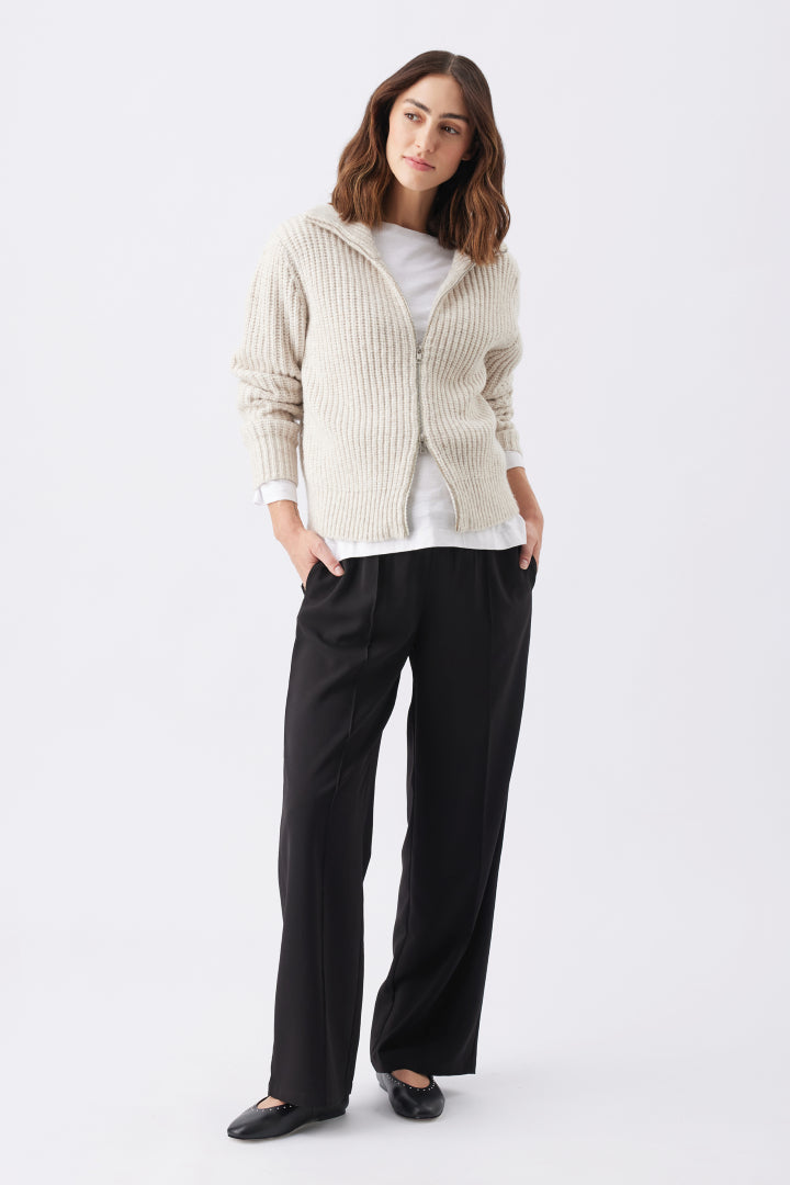 Chunky Zip Knit Maternity Cardigan (Oat) | Ripe Maternity Woman wearing a beige cardigan, white shirt, black pants, and black shoes on a white background