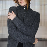 A woman wearing a dark gray heathered knit zip-up sweater with a high mock neck, posed with her arms crossed in front of a modern slatted glass wall.