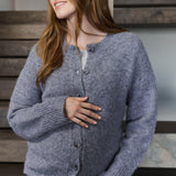 A woman wearing a heathered slate blue-gray knit cardigan sweater with dark buttons, paired with blue jeans, standing in front of a modern wooden wall.