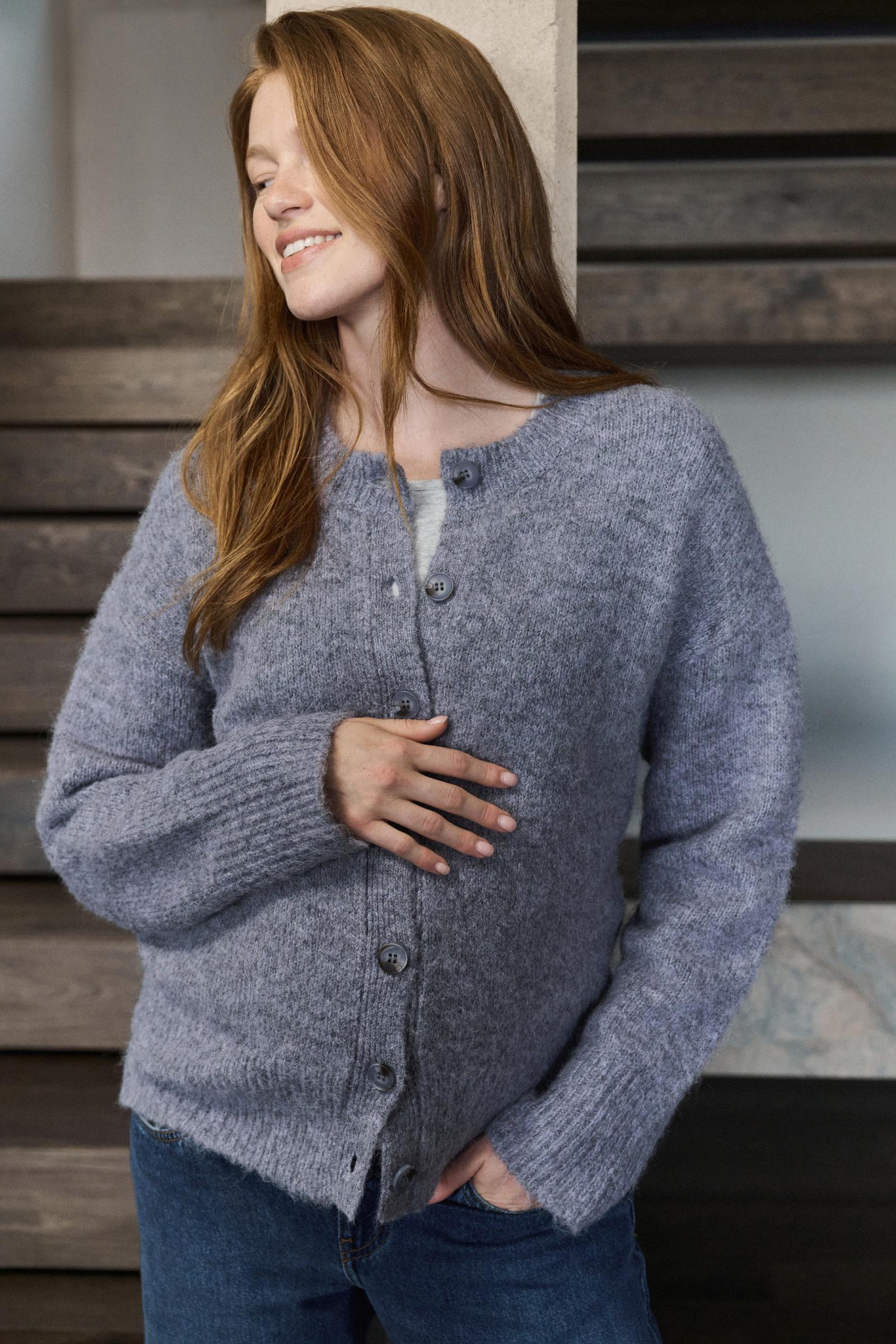 A woman wearing a heathered slate blue-gray knit cardigan sweater with dark buttons, paired with blue jeans, standing in front of a modern wooden wall.