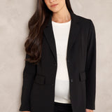 A woman modeling a black tailored blazer over a white top and black pants, standing against a textured light gray wall.