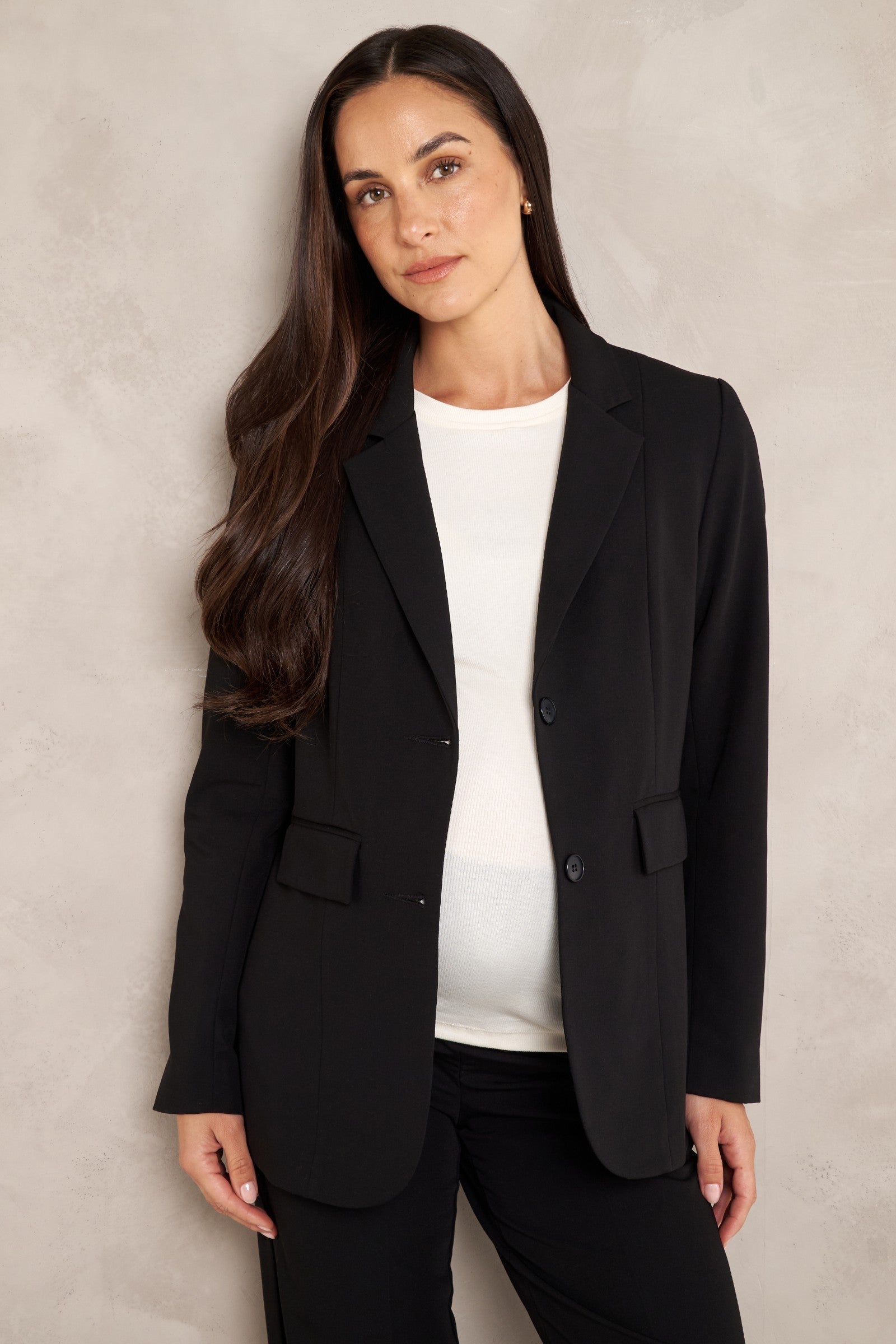 A woman modeling a black tailored blazer over a white top and black pants, standing against a textured light gray wall.