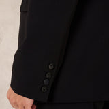 Close-up of a black tailored jacket sleeve with four dark buttons, worn by a model against a light neutral wall.