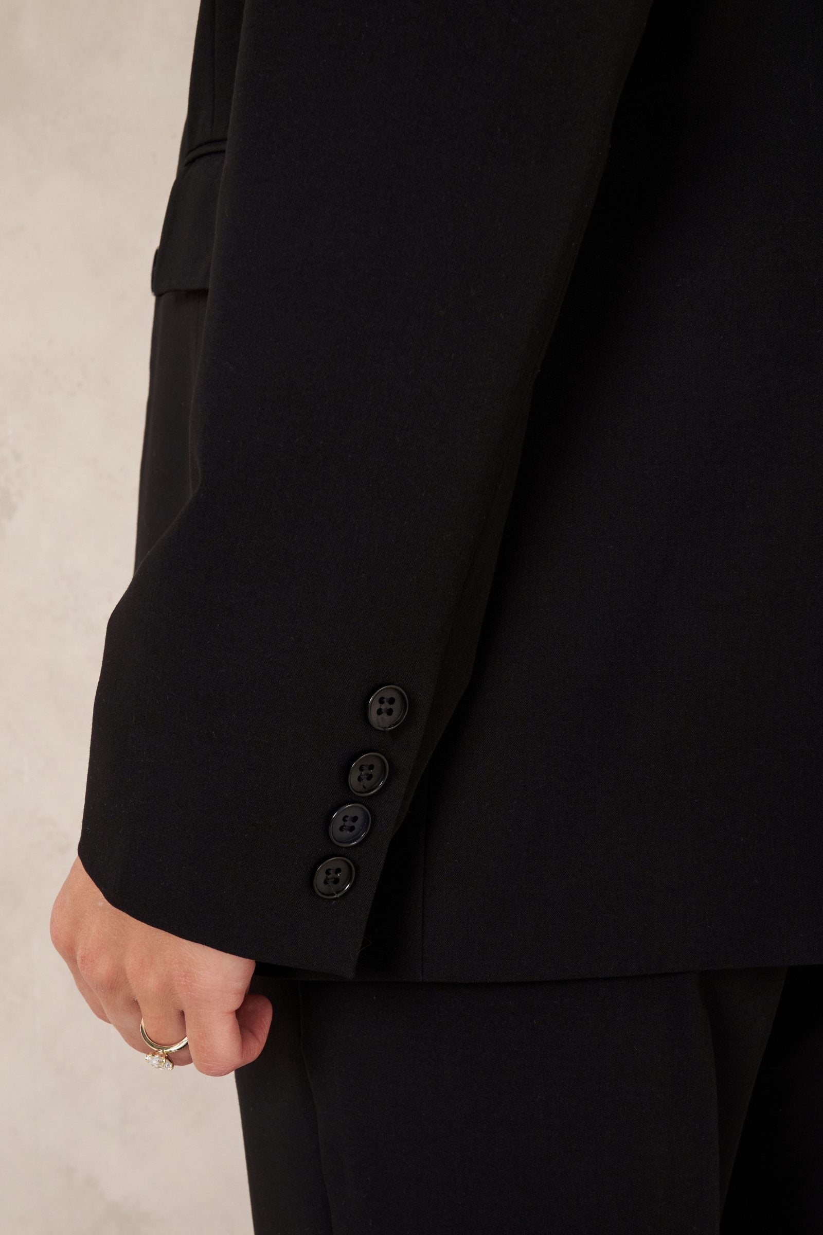 Close-up of a black tailored jacket sleeve with four dark buttons, worn by a model against a light neutral wall.