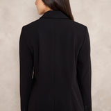A woman shown from the back wearing a black tailored blazer and matching black trousers against a textured light beige wall.