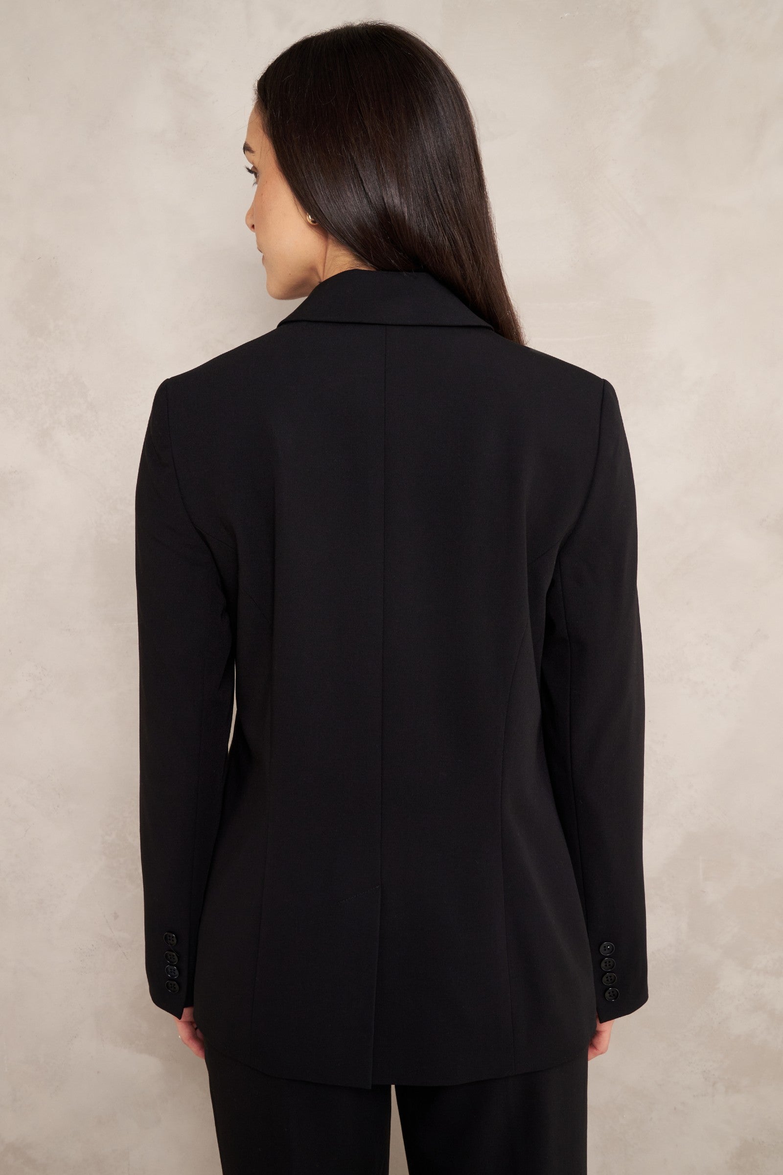 A woman shown from the back wearing a black tailored blazer and matching black trousers against a textured light beige wall.