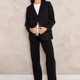 A woman modeling a black tailored blazer and matching wide-leg trousers over a white top, standing against a textured light beige wall.
