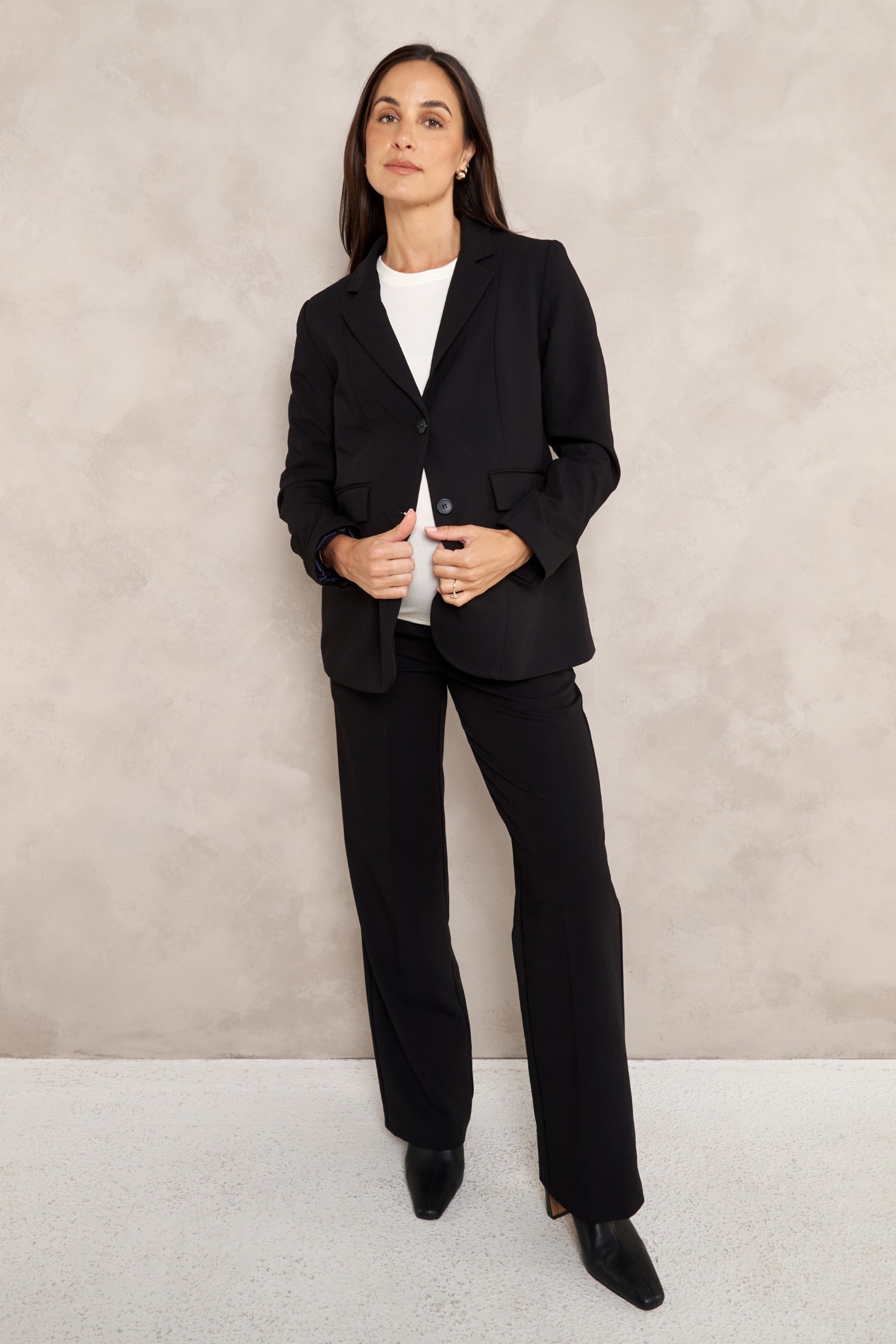 A woman modeling a black tailored blazer and matching wide-leg trousers over a white top, standing against a textured light beige wall.