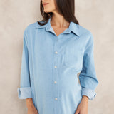 A woman wearing a light blue long-sleeve button-up shirt with rolled cuffs and matching light blue pants, standing against a textured beige wall.