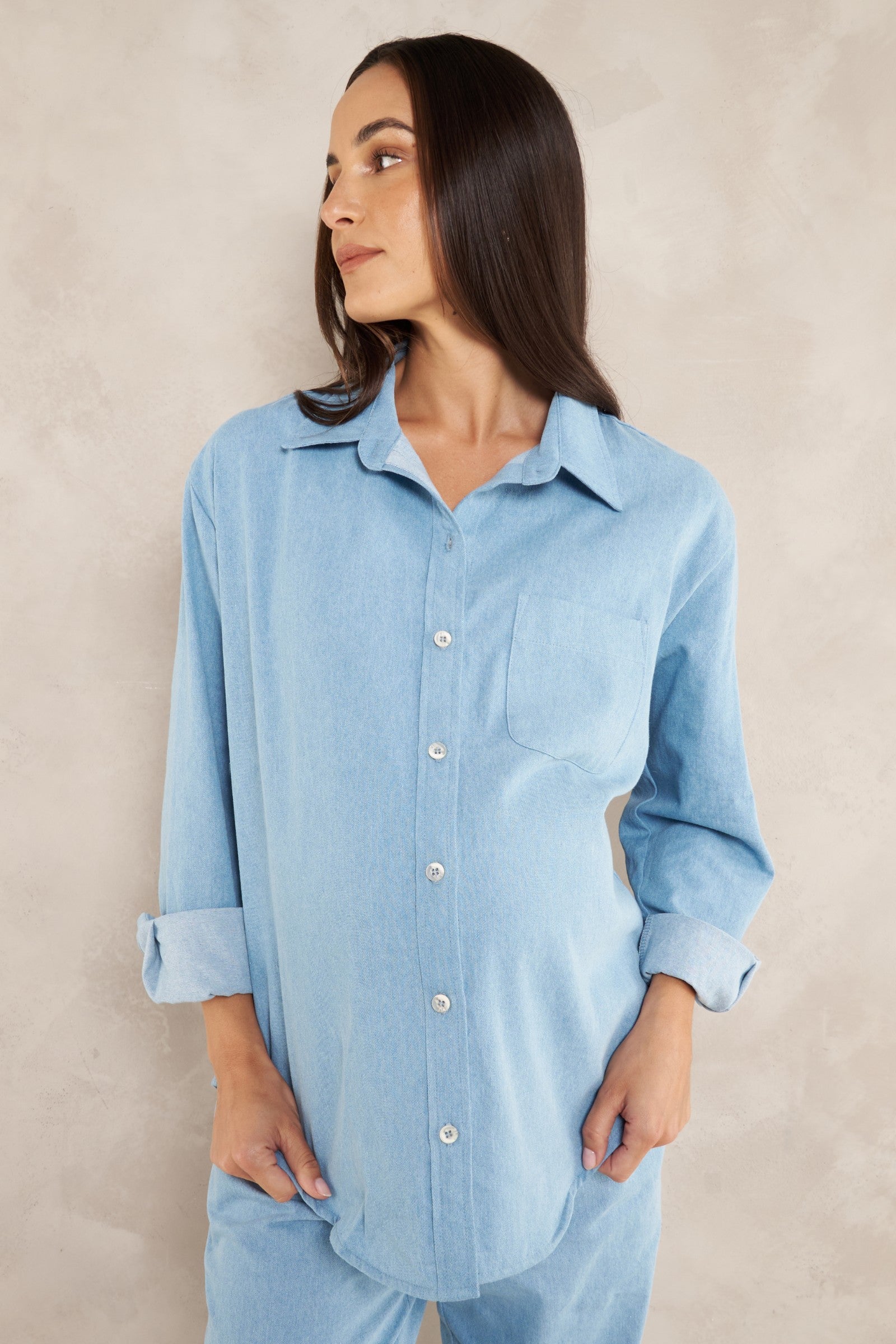A woman wearing a light blue long-sleeve button-up shirt with rolled cuffs and matching light blue pants, standing against a textured beige wall.