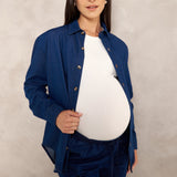 A pregnant woman wearing a navy blue button-up shirt and matching pants over a white top, standing against a textured light gray studio backdrop.