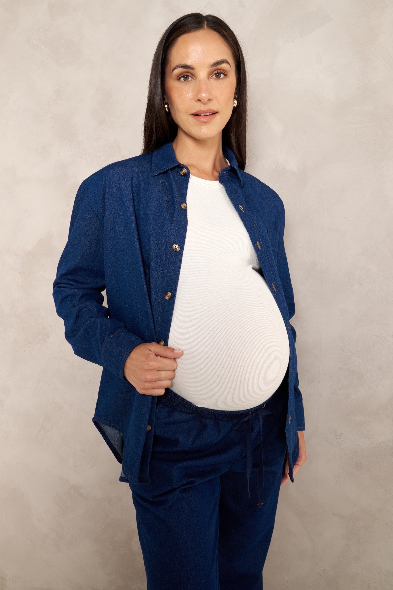 A pregnant woman wearing a navy blue button-up shirt and matching pants over a white top, standing against a textured light gray studio backdrop.
