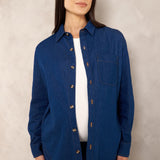 A woman modeling a dark blue button-up shirt with a chest pocket, worn open over a white top and dark pants, standing against a textured light gray wall.