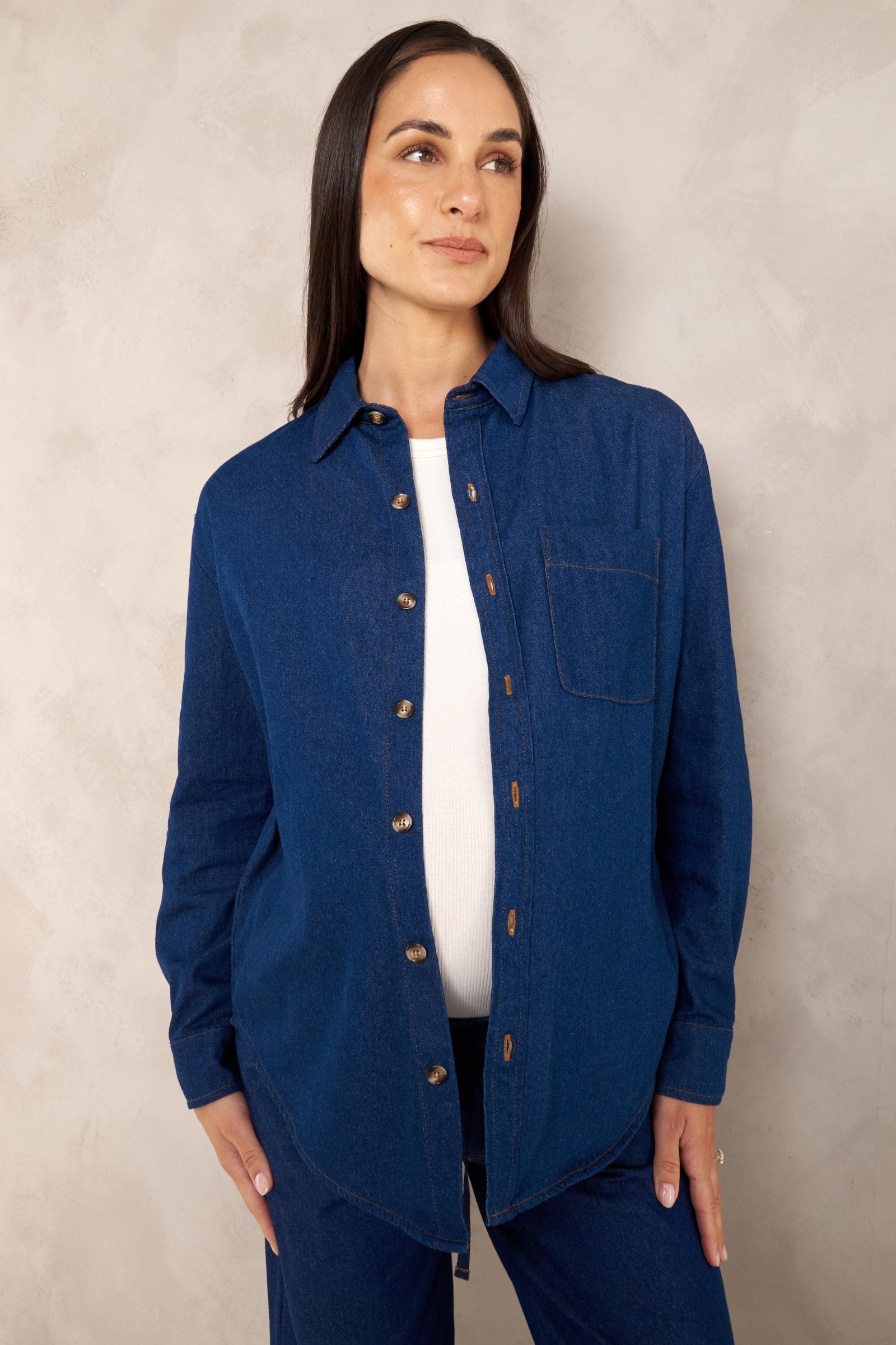 A woman modeling a dark blue button-up shirt with a chest pocket, worn open over a white top and dark pants, standing against a textured light gray wall.
