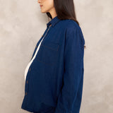 A woman in profile wearing a dark blue denim button-up shirt and matching denim pants, standing against a textured light gray wall.