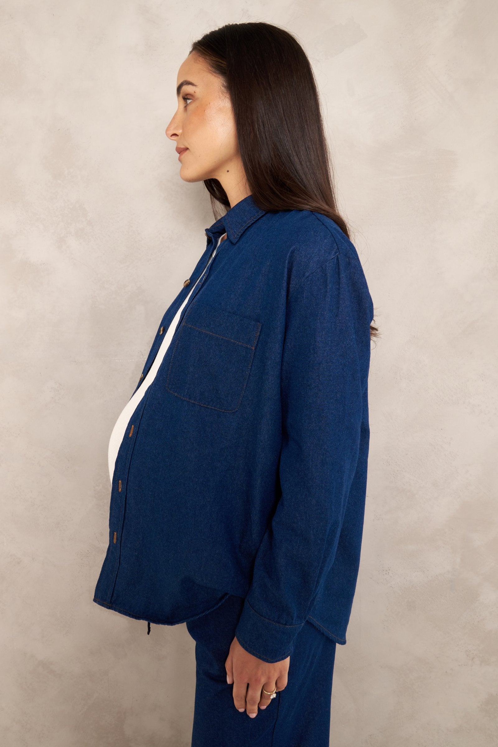 A woman in profile wearing a dark blue denim button-up shirt and matching denim pants, standing against a textured light gray wall.