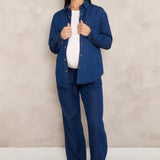 A woman modeling a matching navy blue two-piece set consisting of a long-sleeve button-up shirt and wide-leg trousers, styled over a white top and brown pointed-toe heels, set against a textured beige studio backdrop.