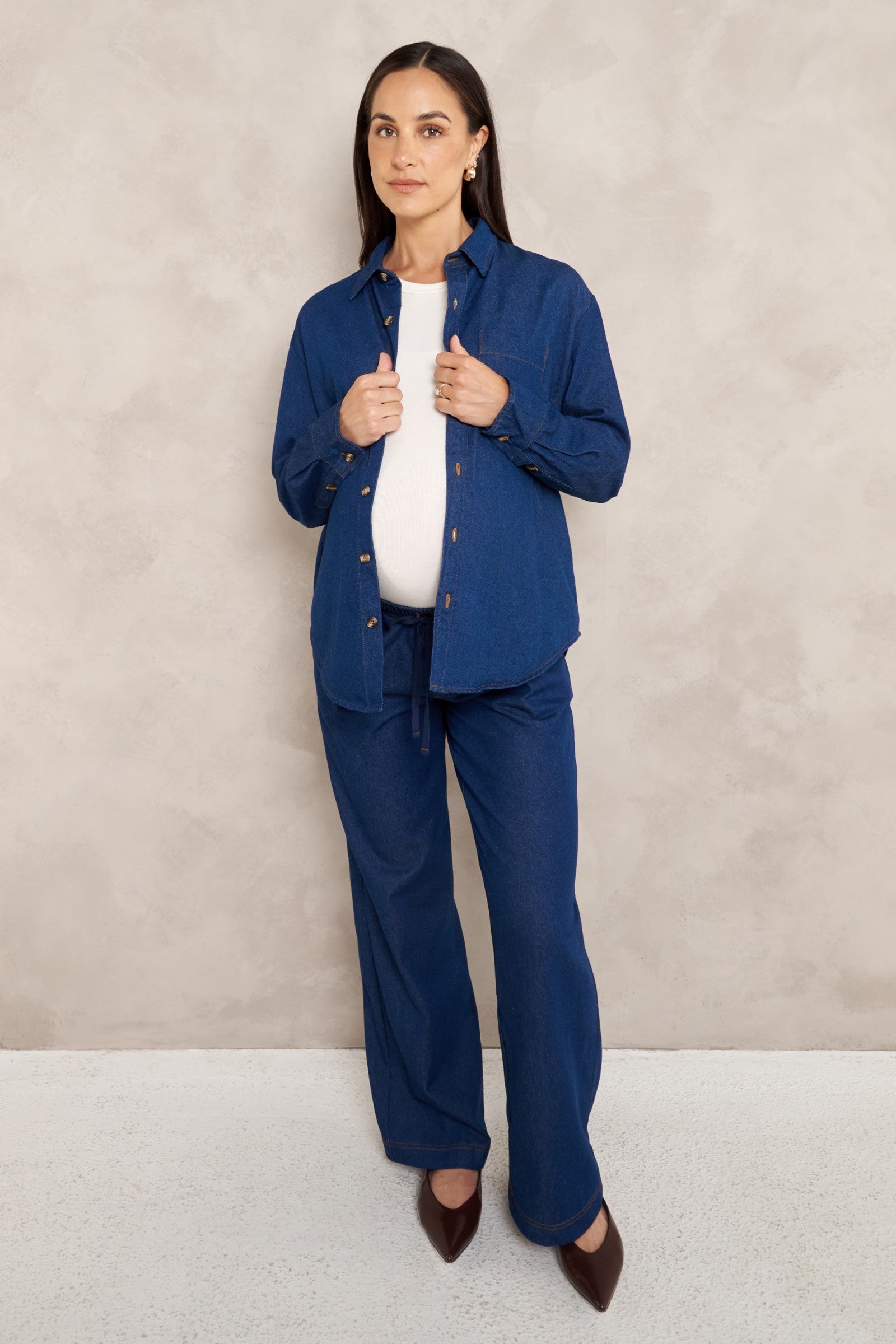 A woman modeling a matching navy blue two-piece set consisting of a long-sleeve button-up shirt and wide-leg trousers, styled over a white top and brown pointed-toe heels, set against a textured beige studio backdrop.