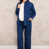 A pregnant model wearing a matching navy blue maternity shirt and pants set over a white top, standing against a textured beige wall.