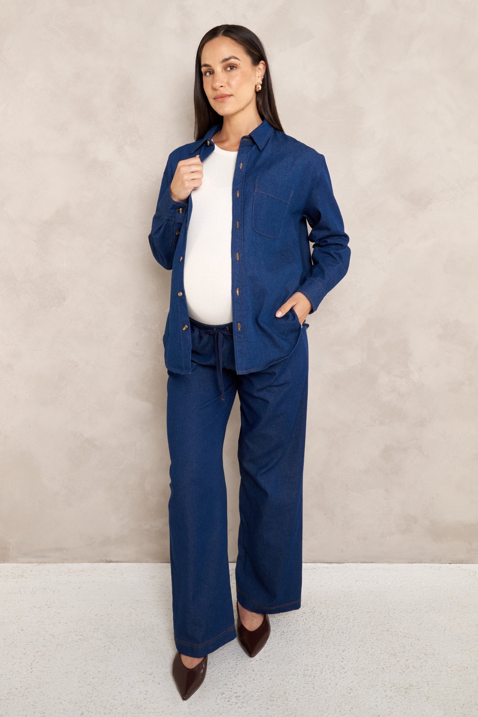 A pregnant model wearing a matching navy blue maternity shirt and pants set over a white top, standing against a textured beige wall.