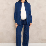 A pregnant woman models a matching navy blue maternity shirt and trouser set with a white top and brown pointed shoes, standing against a textured beige wall.