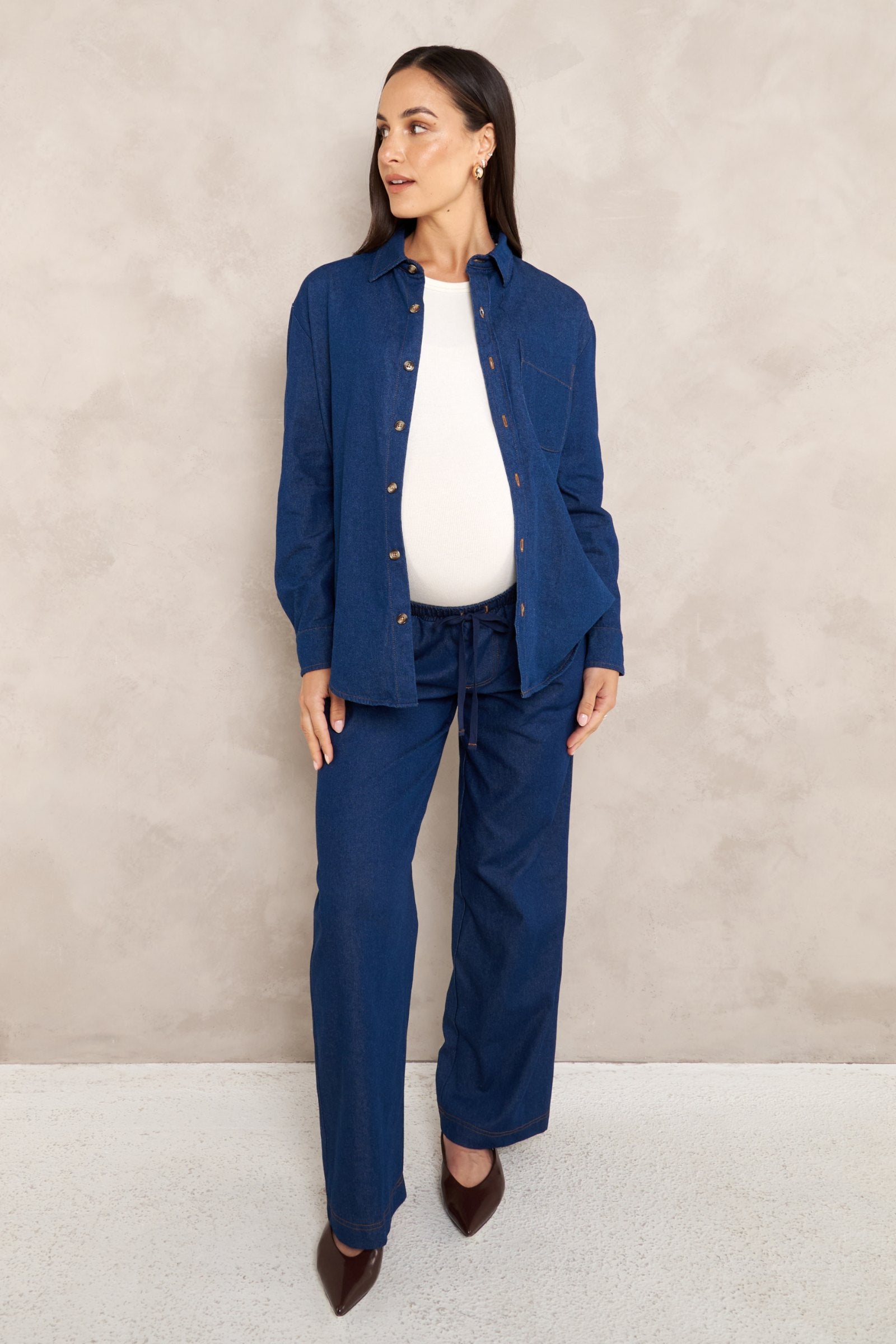 A pregnant woman models a matching navy blue maternity shirt and trouser set with a white top and brown pointed shoes, standing against a textured beige wall.