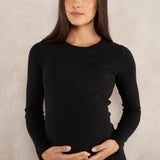 A woman with long dark hair wearing a black long-sleeve crewneck top and blue jeans, standing against a textured light gray background.