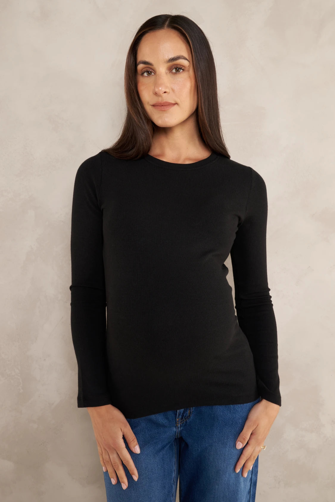 A woman modeling a black long-sleeve crewneck top paired with blue jeans, standing against a textured light gray studio backdrop.