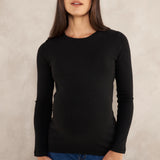 A woman wearing a black long-sleeve crewneck top and blue jeans, standing against a textured light gray studio background.