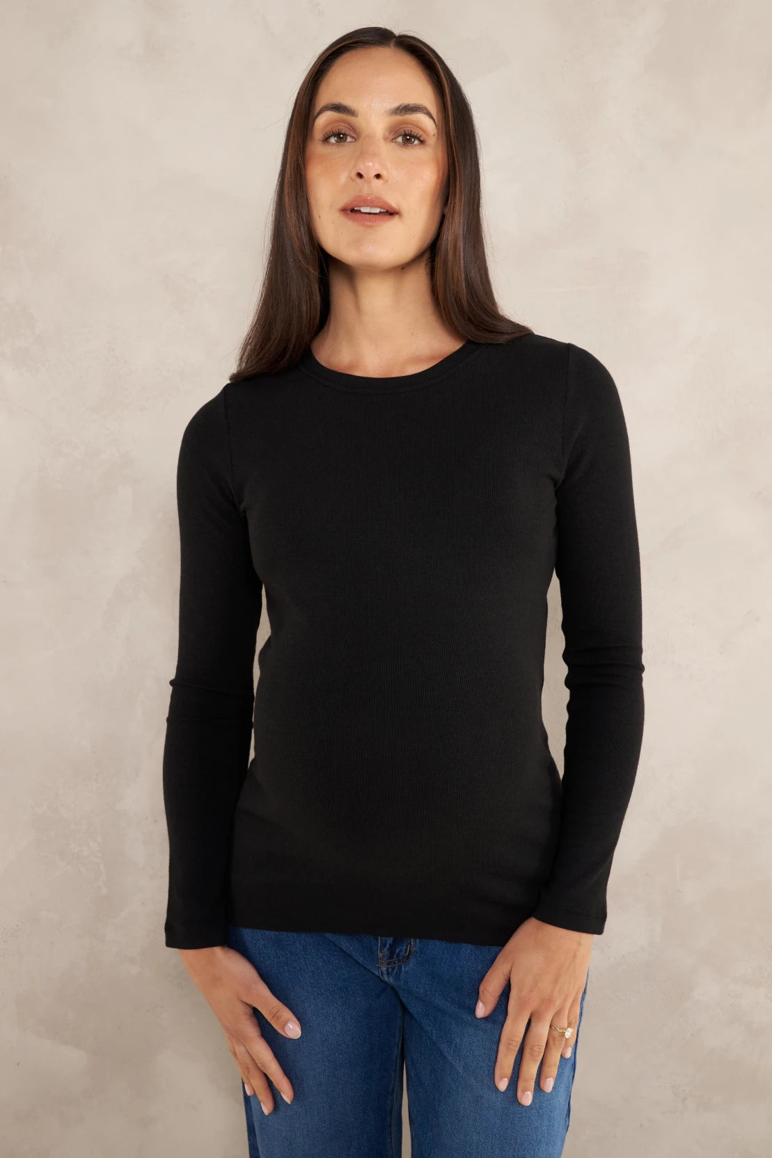 A woman wearing a black long-sleeve crewneck top and blue jeans, standing against a textured light gray studio background.