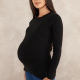 A pregnant woman wearing a black long-sleeve crewneck maternity top and blue jeans, standing against a textured light beige studio backdrop.