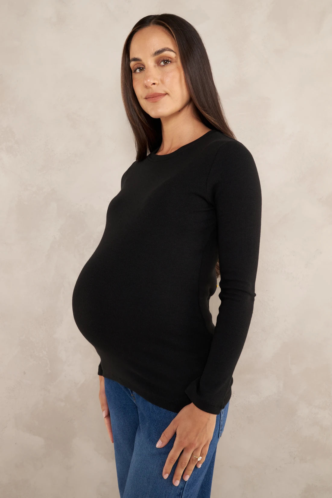 A pregnant woman wearing a black long-sleeve crewneck maternity top and blue jeans, standing against a textured light beige studio backdrop.