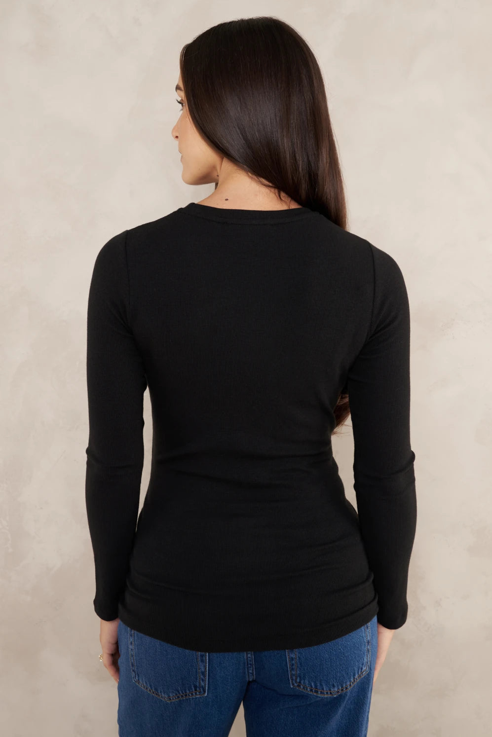 A woman viewed from the back wearing a fitted black long-sleeve top and blue jeans, standing against a textured light beige wall.
