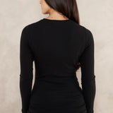 A woman viewed from the back wearing a fitted black long-sleeve top and blue jeans, standing against a textured light beige wall.