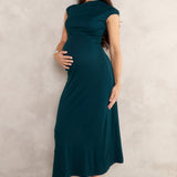 A pregnant woman wearing an emerald green maternity maxi dress with cap sleeves and black pointed-toe heels, standing against a textured light beige wall.
