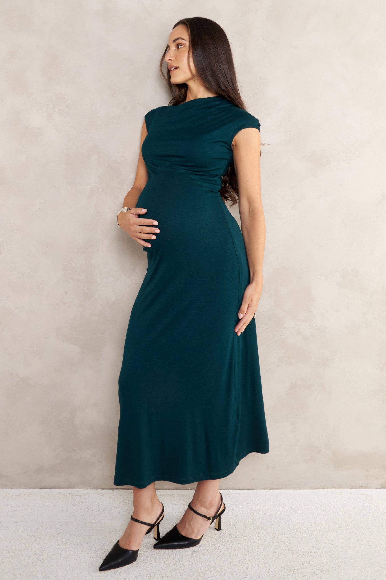A pregnant woman wearing an emerald green maternity maxi dress with cap sleeves and black pointed-toe heels, standing against a textured light beige wall.