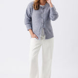 A woman modeling a light blue-gray fuzzy knit cardigan sweater with white wide-leg pants and brown shoes, standing against a plain white studio background.