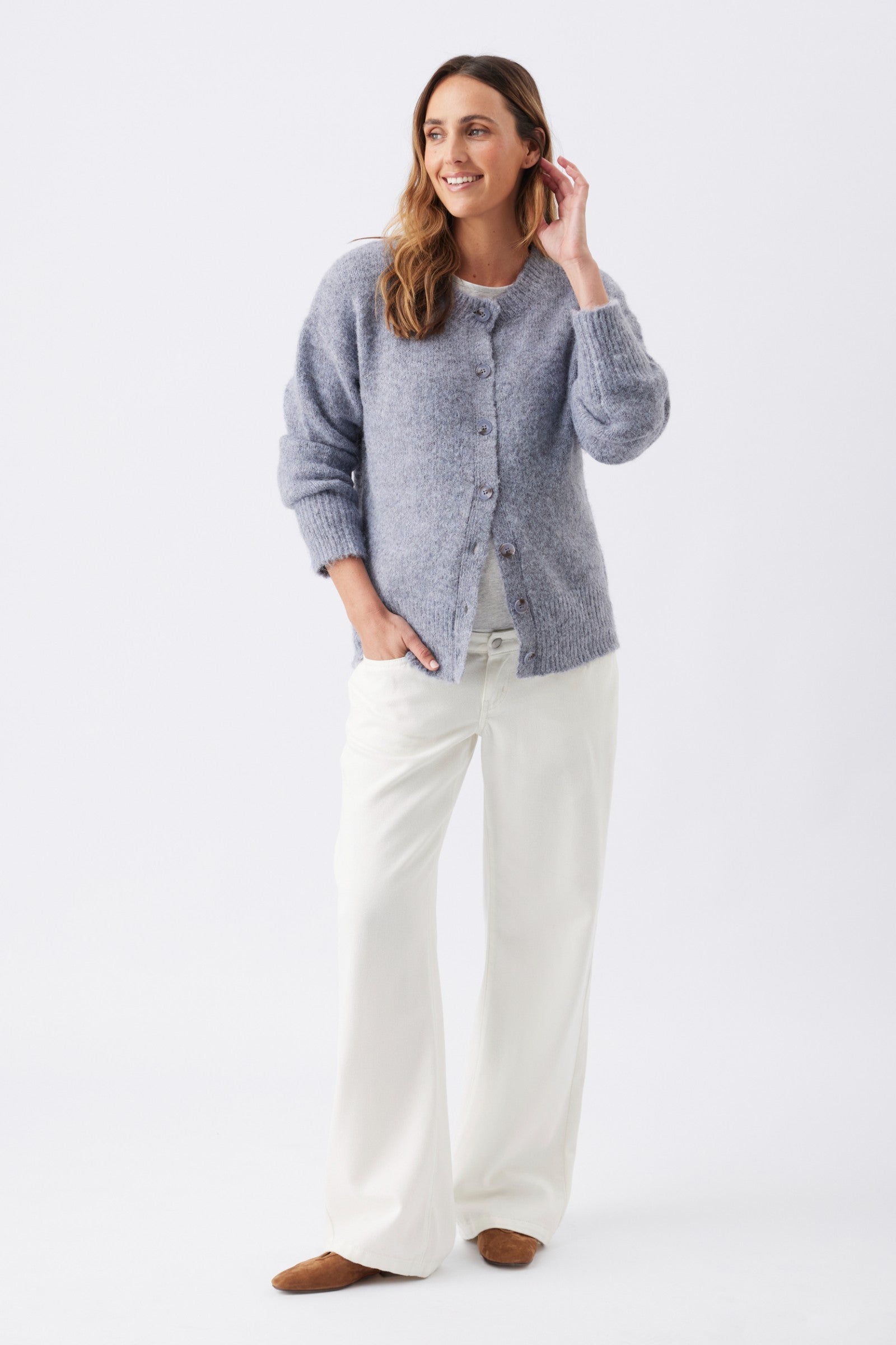 A woman modeling a light blue-gray fuzzy knit cardigan sweater with white wide-leg pants and brown shoes, standing against a plain white studio background.