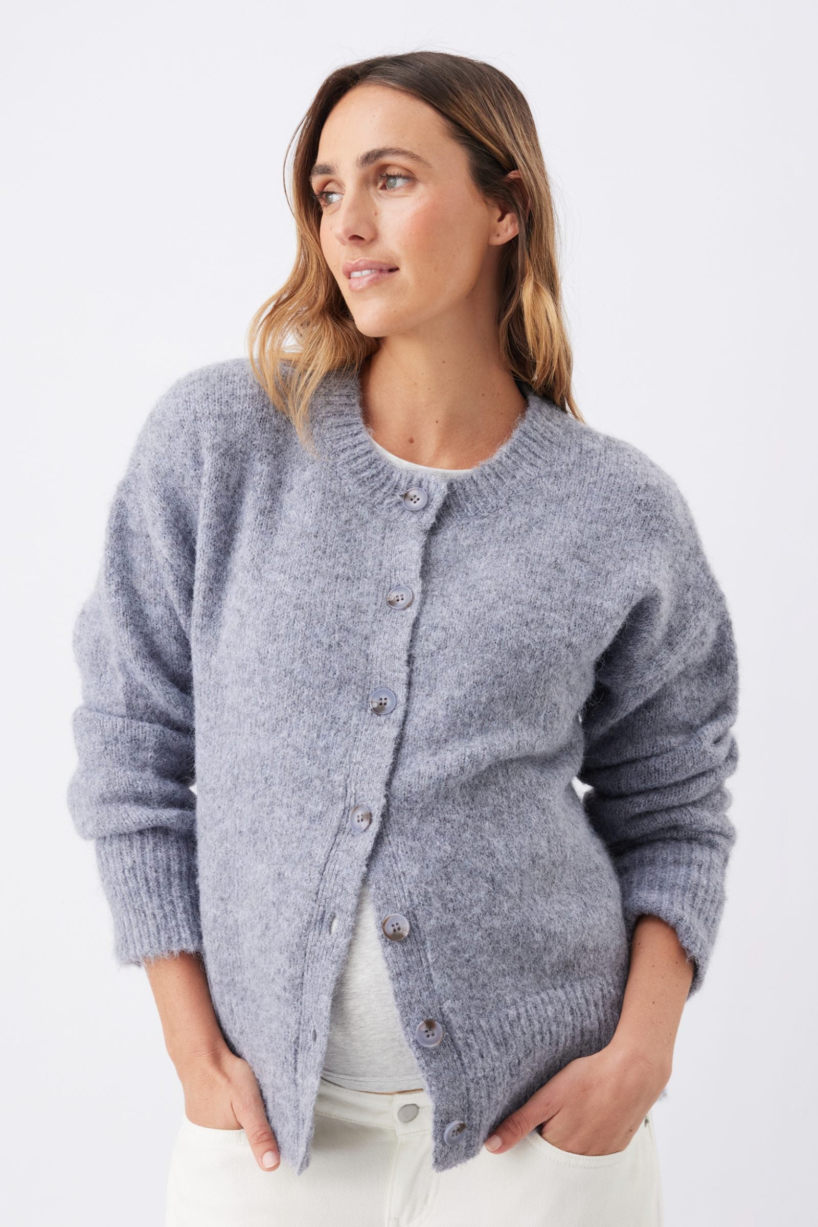 A woman wearing a light gray heathered knit cardigan with a button-up front, styled over a white top and white pants, photographed against a clean white studio background.