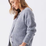 A woman wearing a fuzzy light gray cardigan sweater with white pants, photographed against a clean light gray studio background.