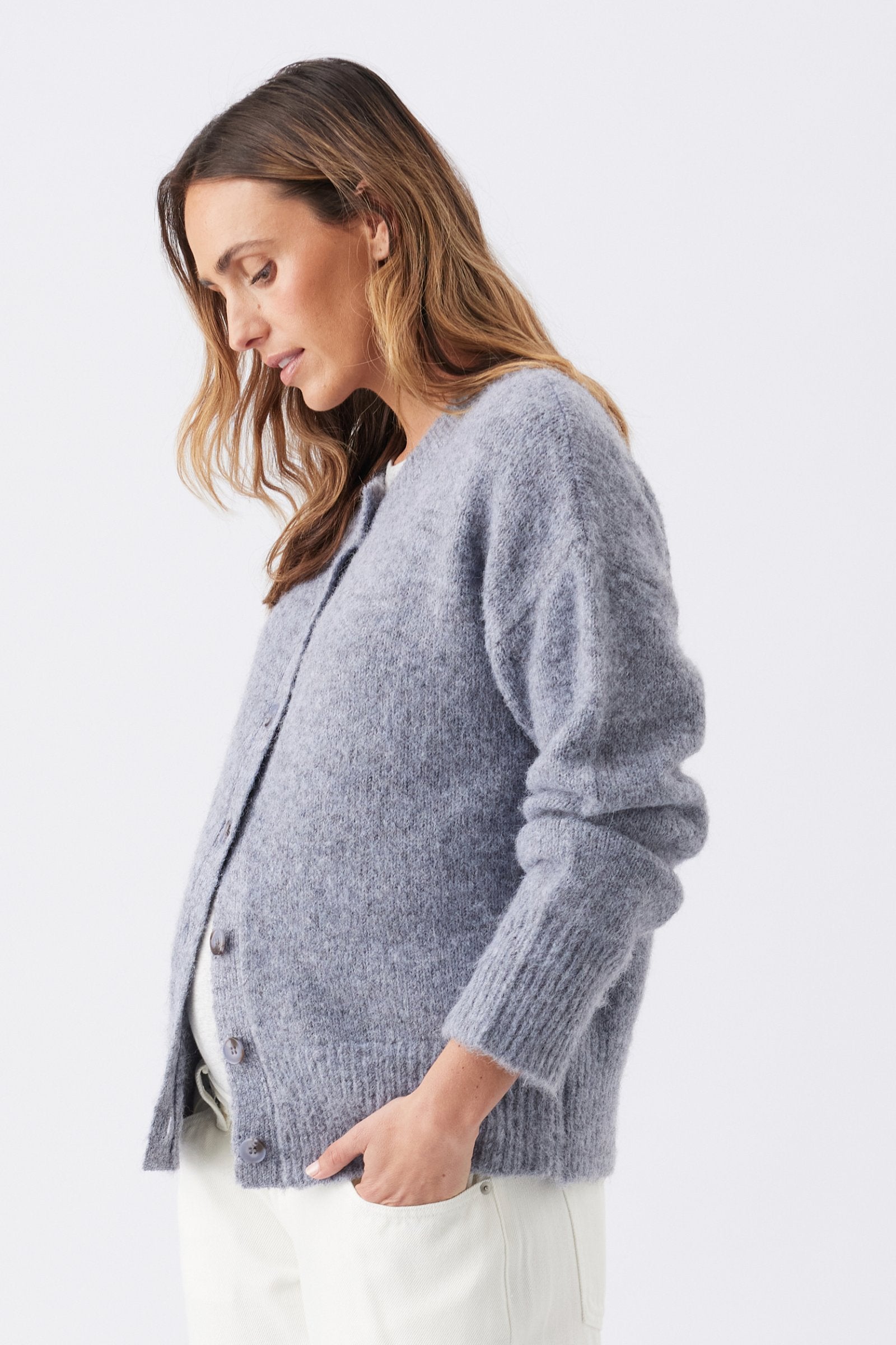 A woman wearing a fuzzy light gray cardigan sweater with white pants, photographed against a clean light gray studio background.