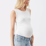 A pregnant woman wearing a white sleeveless maternity tank top and light blue jeans, standing against a plain white studio background.