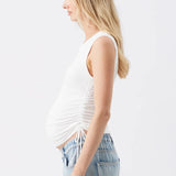 A pregnant woman shown in profile wearing a white ruched tank top and light blue jeans against a plain white studio background.