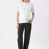 A woman modeling a white short-sleeve top with a layered hem and dark charcoal wide-leg pants, standing against a plain white studio background.