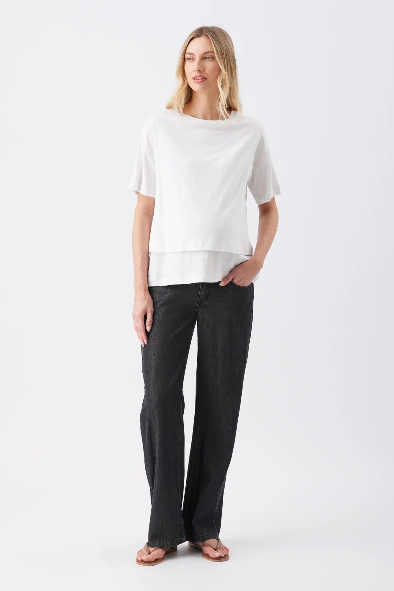 A woman modeling a white short-sleeve top with a layered hem and dark charcoal wide-leg pants, standing against a plain white studio background.