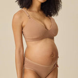 A pregnant model wearing a matching beige maternity bra and underwear set, standing against a solid beige background.