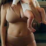 A woman wearing a matching beige nursing bra and underwear holds a baby dressed in a white long-sleeve outfit in a kitchen setting.