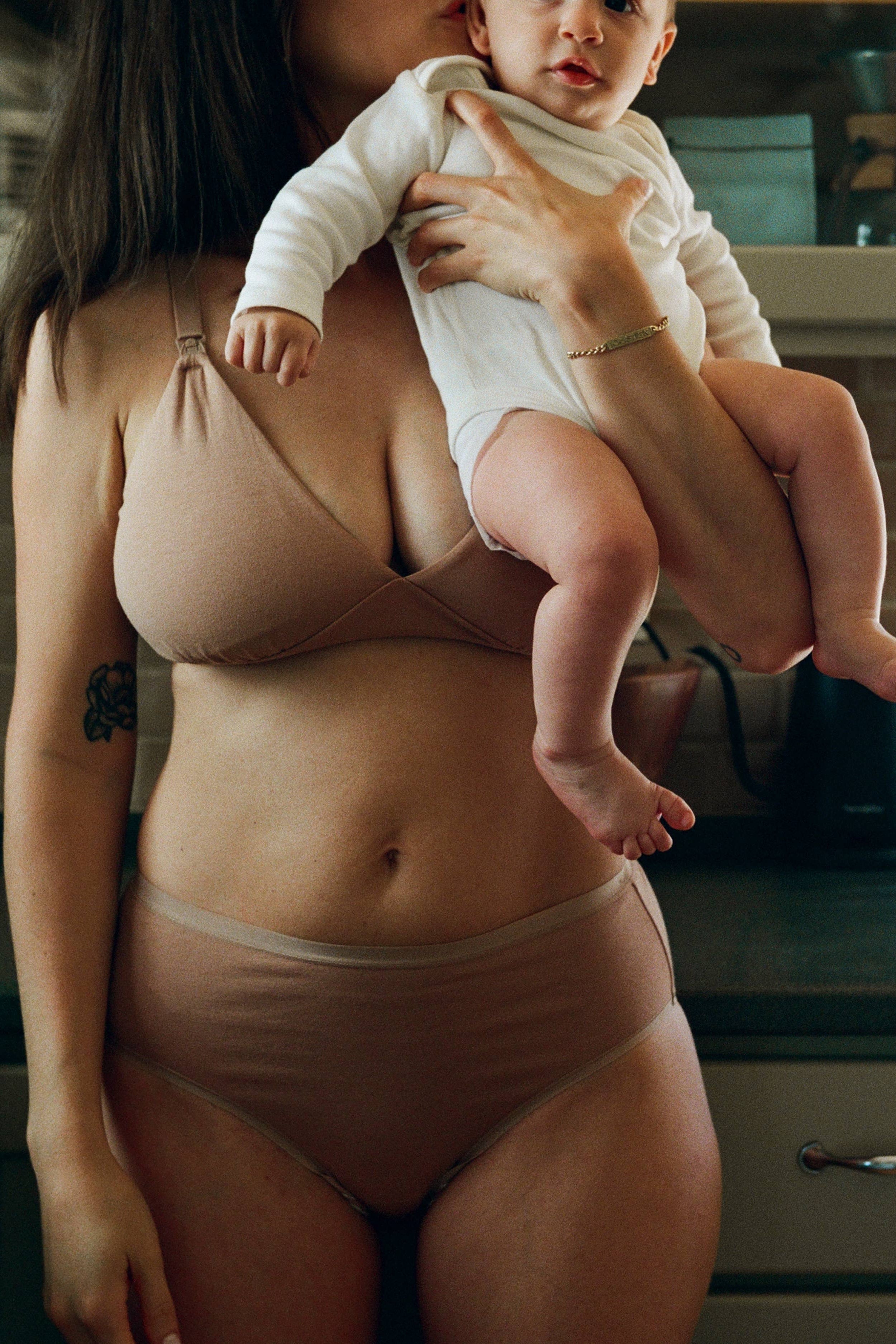 A woman wearing a matching beige nursing bra and underwear holds a baby dressed in a white long-sleeve outfit in a kitchen setting.