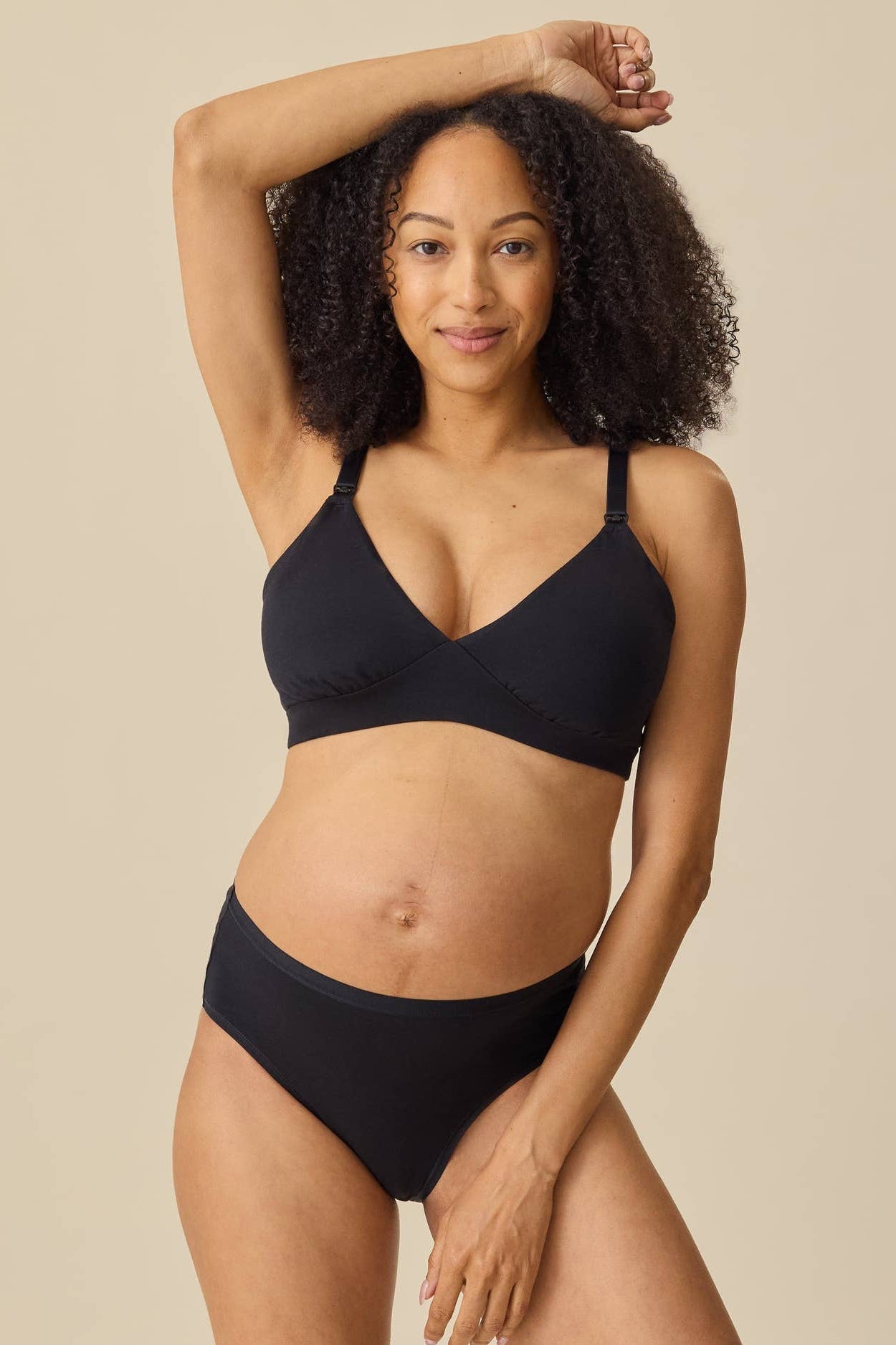 A pregnant model wearing a black maternity bra and matching black underwear, posing against a solid beige background.